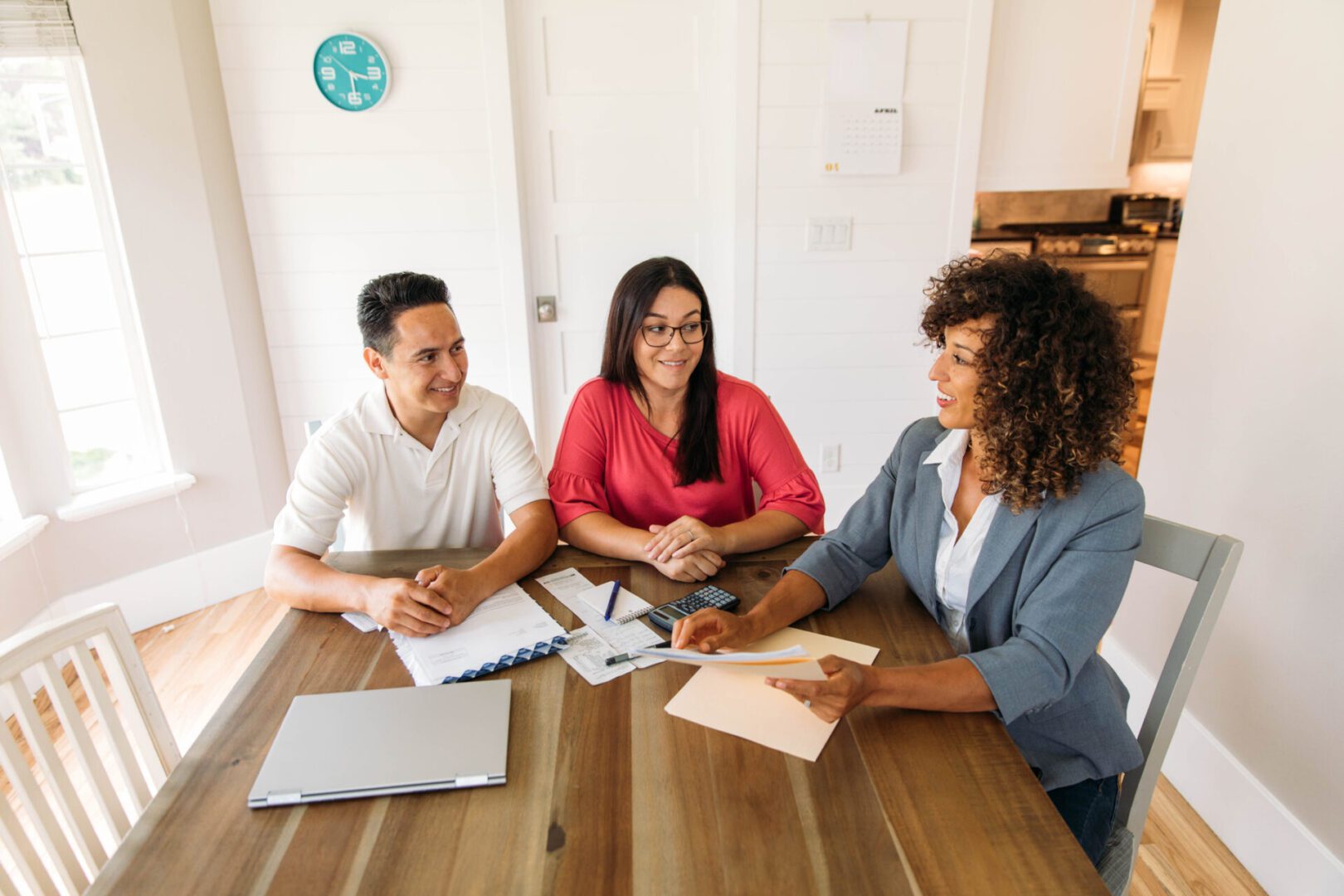 A female financial advisor sits with a millennial age married couple at a dining room table with laptop and financial reports helping with their monthly budget and investments. She is showing her clients how to read financial reports and monthly finances, pay taxes and save money for the future.